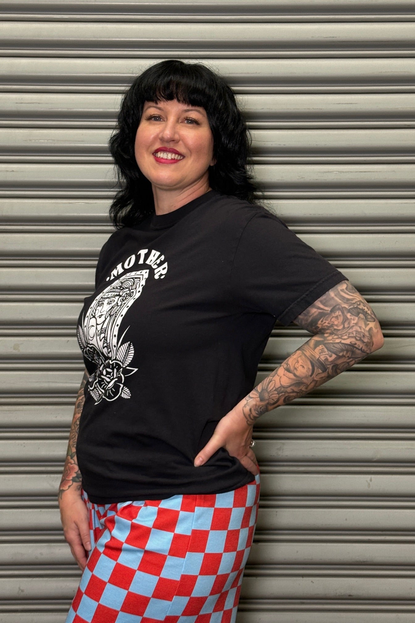 Person wearing a black t-shirt with graphic design and a red and blue checkered skirt, standing against a metallic shutter.