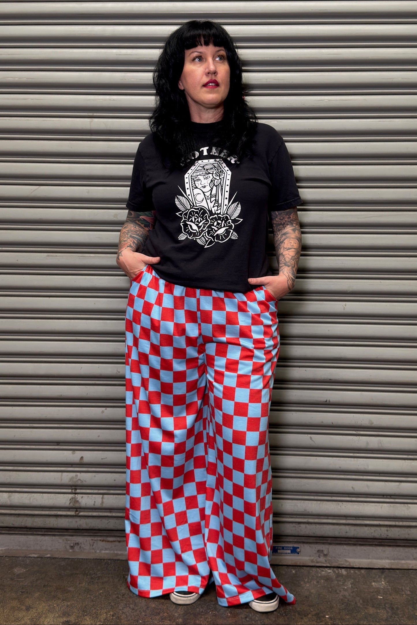 Person wearing a black t-shirt with a graphic design and red and blue checkered pants standing against a metal shutter.