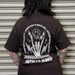 Person wearing a black t-shirt with a graphic design and text on the back, standing in front of a closed metal shutter.