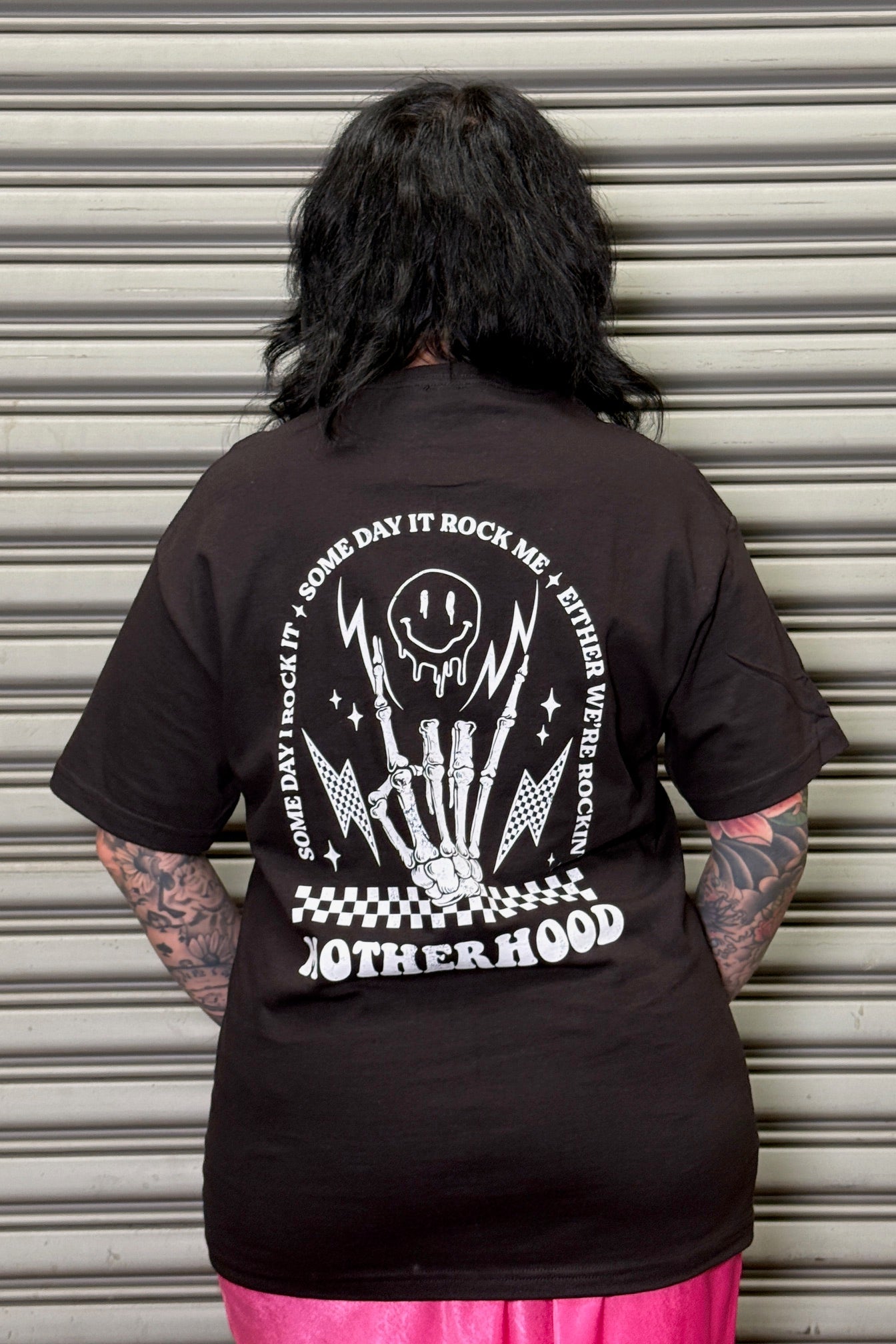 Person wearing a black t-shirt with a graphic design and text on the back, standing in front of a closed metal shutter.