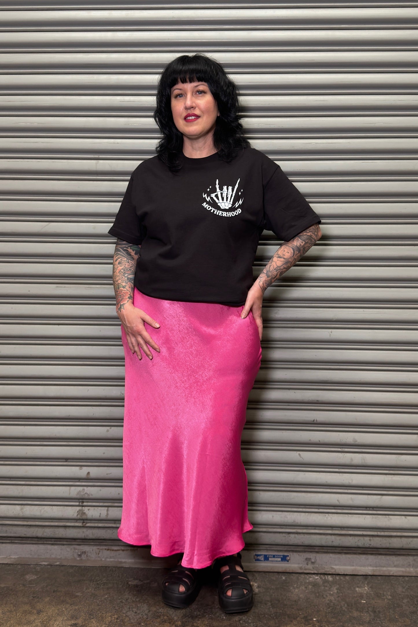 Person wearing a black t-shirt and pink skirt standing in front of a metal shutter.