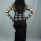 A woman with long black hair is shown from behind, wearing a white and black plaid long-sleeve top with sheer mesh sleeves and black wide-leg pants, posing against a light gray studio backdrop.