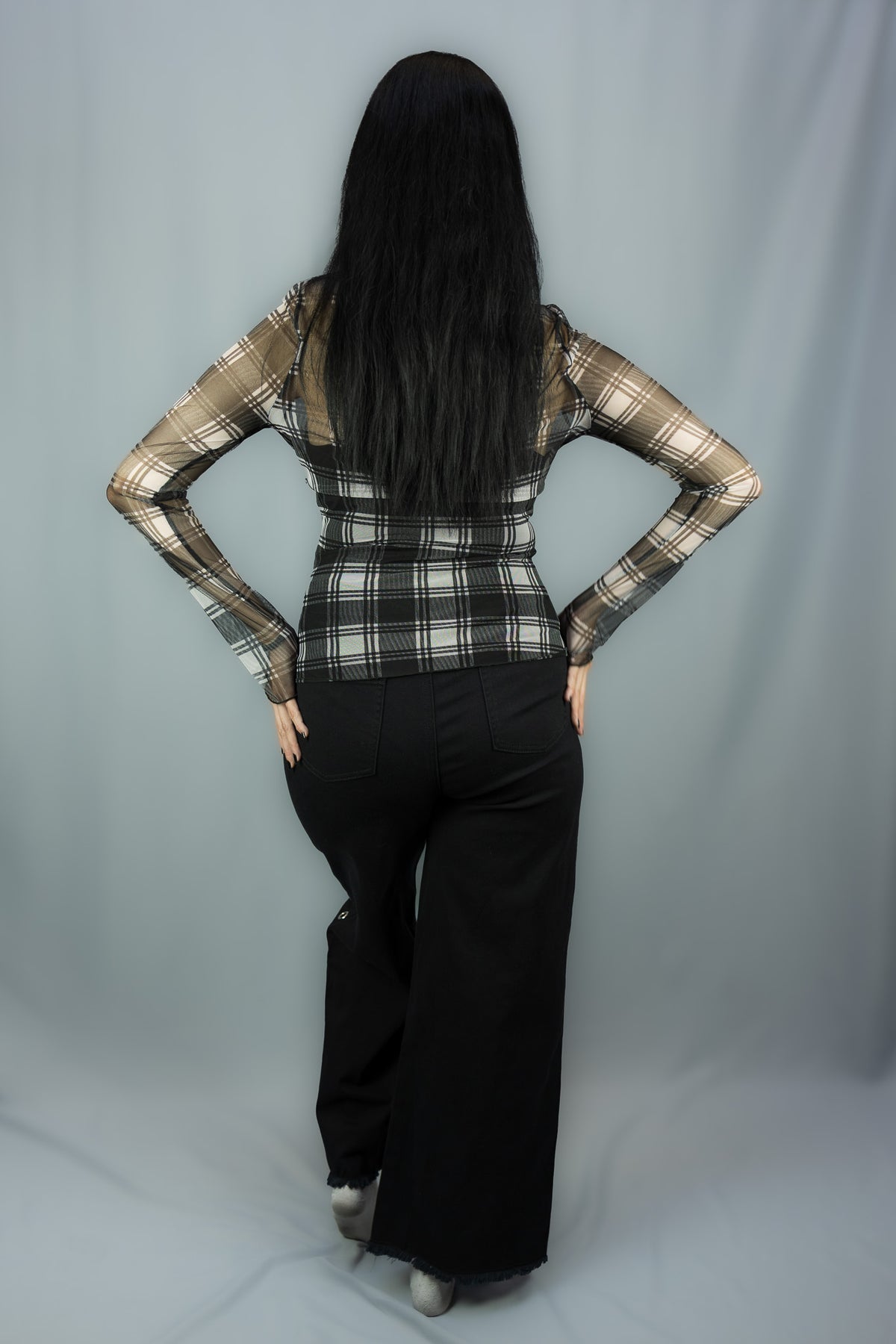 A woman with long black hair is shown from behind, wearing a white and black plaid long-sleeve top with sheer mesh sleeves and black wide-leg pants, posing against a light gray studio backdrop.
