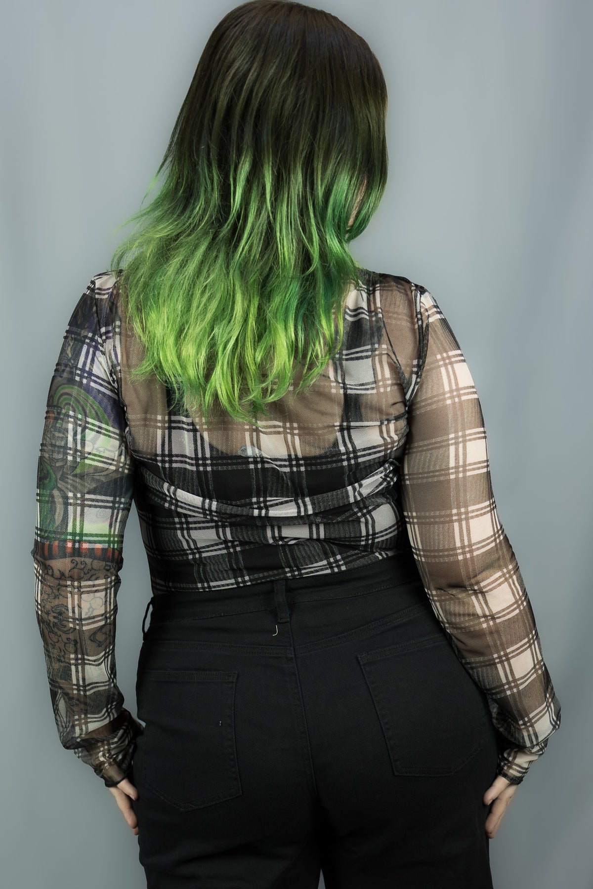 A person shown from the back wearing a sheer long-sleeve plaid top with black and white tones and bright green ombre hair, paired with black pants, against a plain gray background.