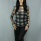 A woman modeling a sheer long-sleeve top with a black and white plaid pattern over a black tank top, paired with black pants featuring eyelet details down the sides, set against a plain gray studio backdrop.