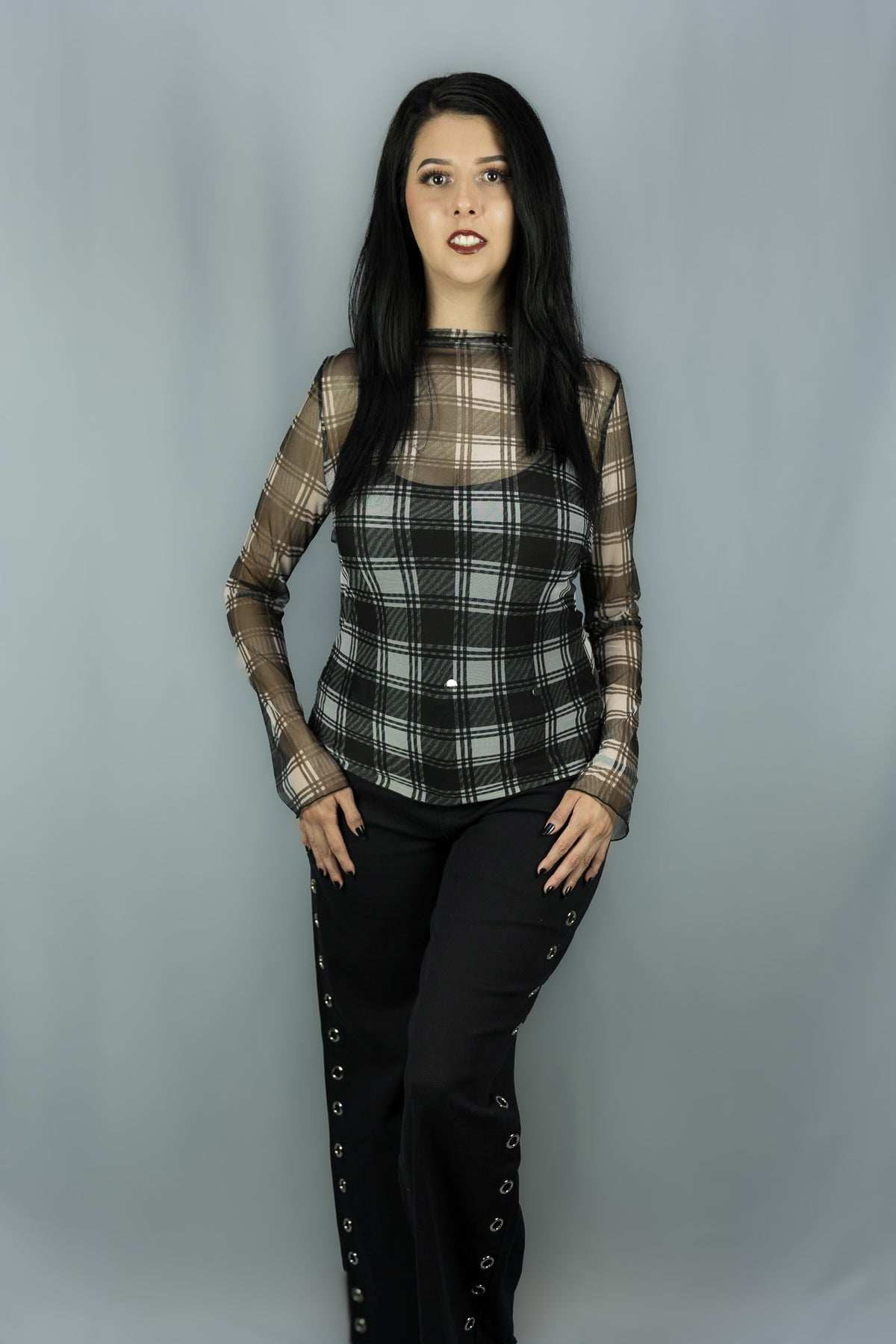 A woman modeling a sheer long-sleeve top with a black and white plaid pattern over a black tank top, paired with black pants featuring eyelet details down the sides, set against a plain gray studio backdrop.