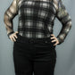 A person wearing a sheer long-sleeve plaid top in black and white paired with black pants, standing against a plain gray backdrop.