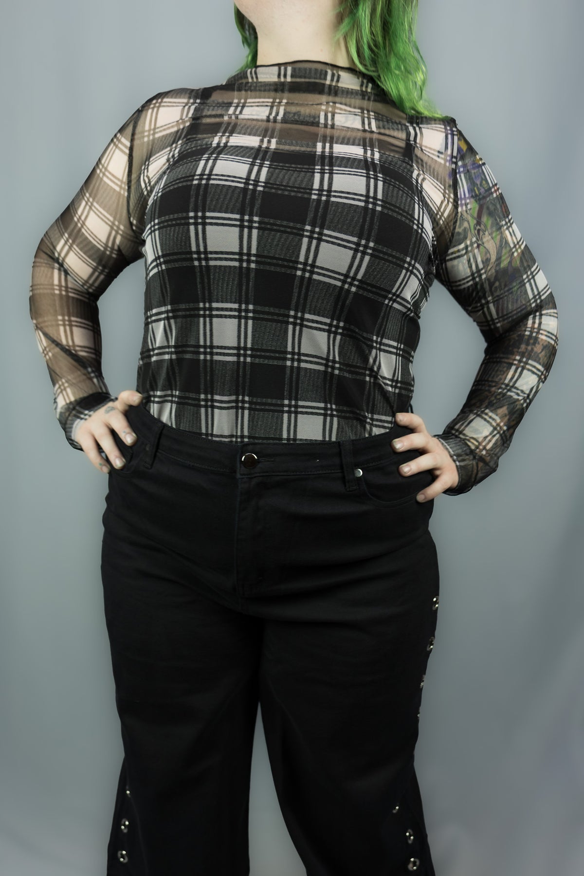 A person wearing a sheer long-sleeve plaid top in black and white paired with black pants, standing against a plain gray backdrop.