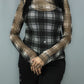 A woman wearing a sheer long-sleeve mesh top with a black and white plaid pattern over a black camisole, paired with black pants, set against a plain gray background.