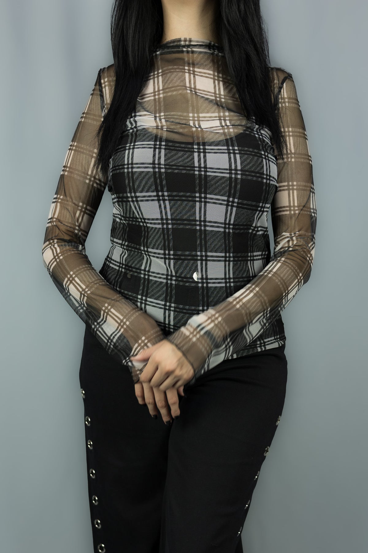A woman wearing a sheer long-sleeve mesh top with a black and white plaid pattern over a black camisole, paired with black pants, set against a plain gray background.