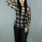 A woman modeling a sheer long-sleeve plaid top in black and white paired with black pants featuring side grommets, against a solid gray studio backdrop.