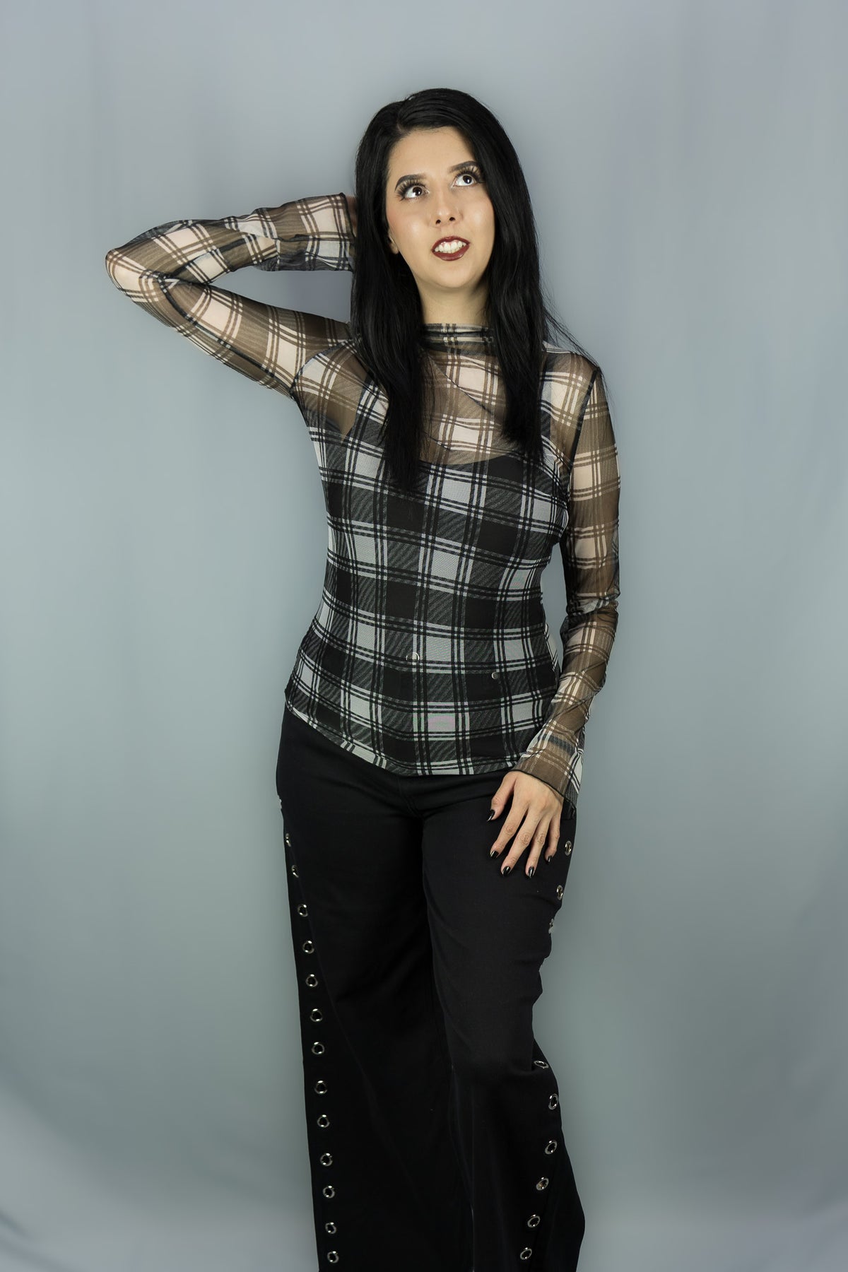 A woman modeling a sheer long-sleeve plaid top in black and white paired with black pants featuring side grommets, against a solid gray studio backdrop.
