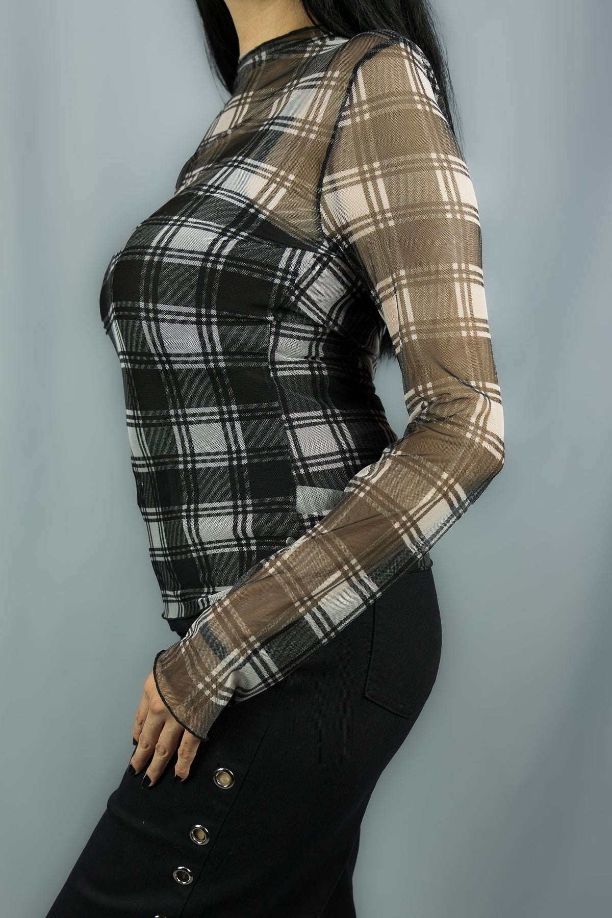A woman wearing a sheer long-sleeve mesh top with a black and white plaid pattern, paired with black pants, shown from a side profile against a solid gray background.