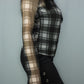 A woman wearing a sheer long-sleeve plaid top in black and white, paired with black wide-leg pant featuring decorative side buttons, set against a plain gray background.