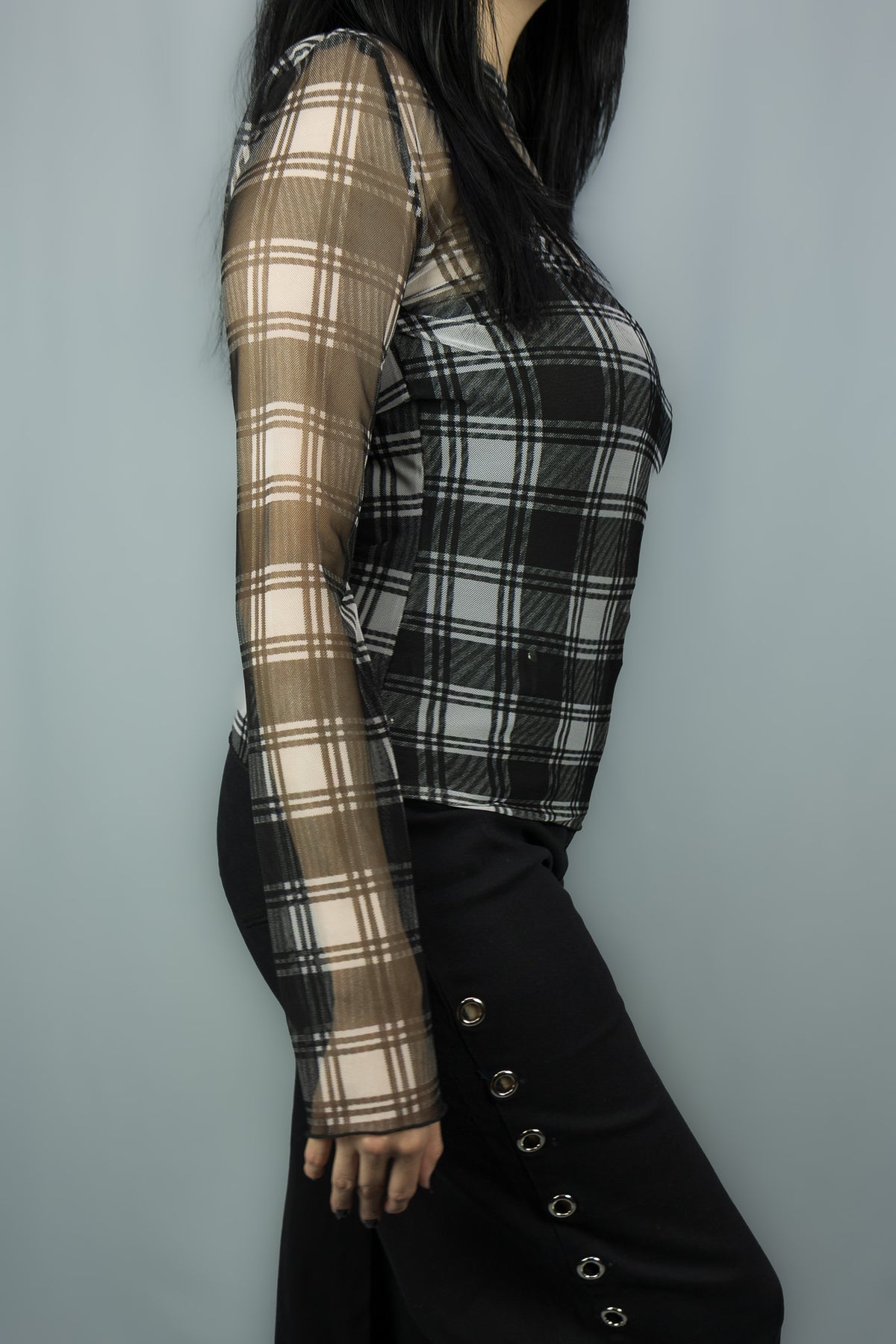 A woman wearing a sheer long-sleeve plaid top in black and white, paired with black wide-leg pant featuring decorative side buttons, set against a plain gray background.