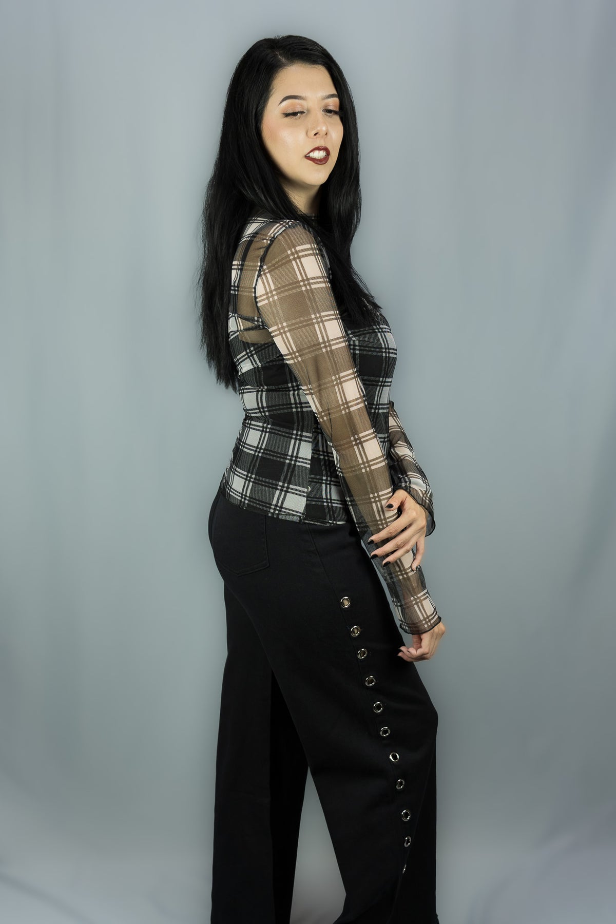 A woman modeling a long-sleeve sheer mesh top with a black and white plaid pattern, paired with black wide-leg pants featuring decorative buttons down the side, set against a plain gray studio backdrop.