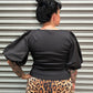 A person wearing a black long-sleeve top and a leopard print skirt, photographed from above against a corrugated metal wall.