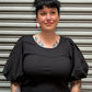A woman with short dark hair and visible tattoos is shown from the side against a corrugated metal shutter. She is wearing a black ribbed top and a leopard-print skirt.