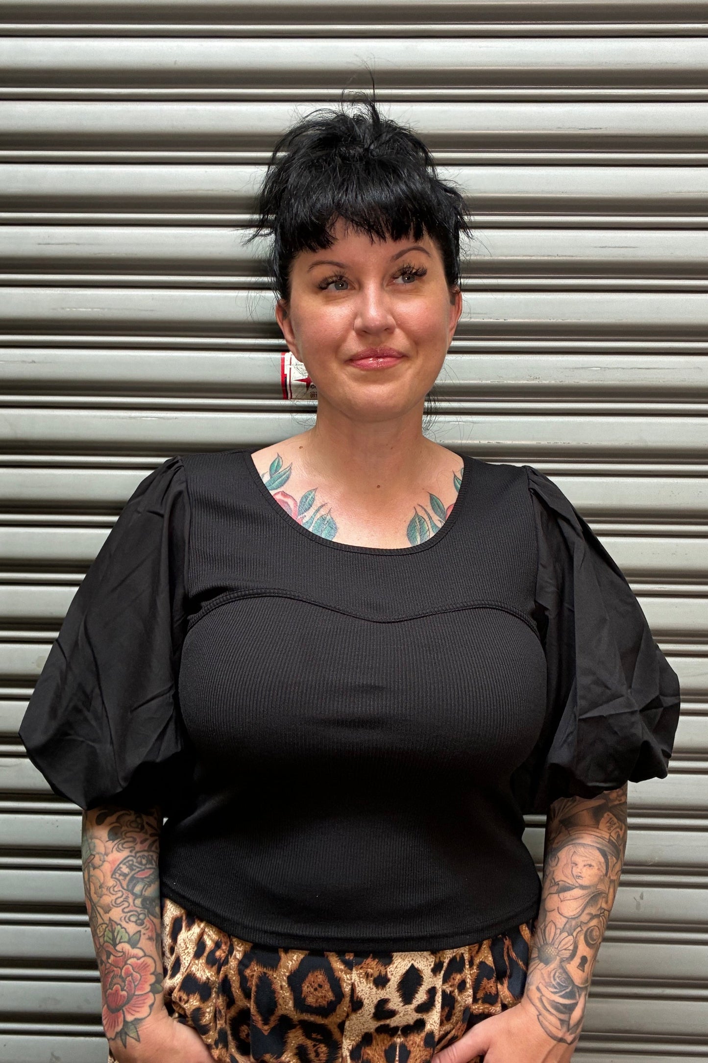 A woman with short dark hair and visible tattoos is shown from the side against a corrugated metal shutter. She is wearing a black ribbed top and a leopard-print skirt.