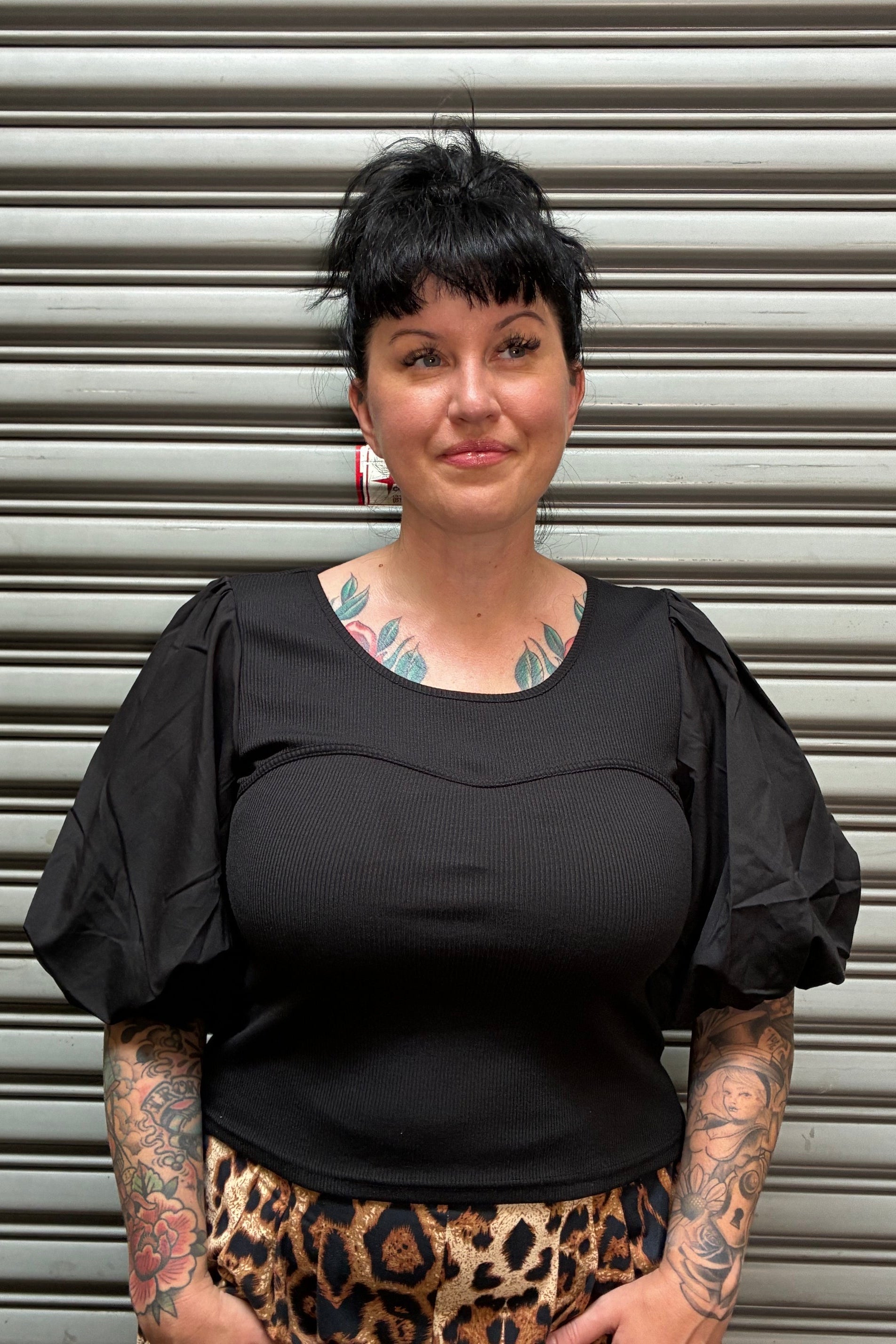 A woman with short dark hair and visible tattoos is shown from the side against a corrugated metal shutter. She is wearing a black ribbed top and a leopard-print skirt.