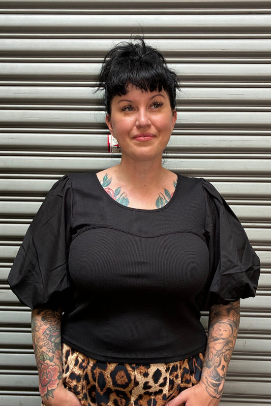 A woman with short dark hair and visible tattoos is shown from the side against a corrugated metal shutter. She is wearing a black ribbed top and a leopard-print skirt.