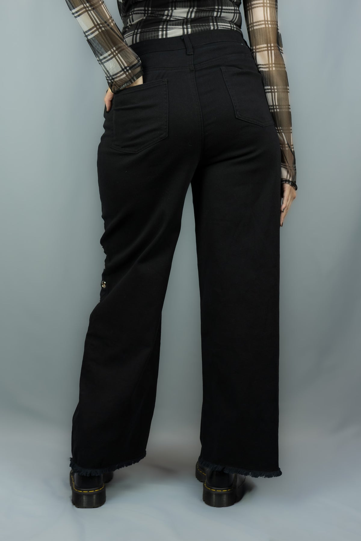A person shown from the back wearing black wide-leg pants with a frayed hem, paired with a white and black plaid long-sleeve shirt and black platform shoes, against a gray studio backdrop.