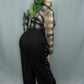 A model with green-tipped hair is shown from a rear three-quarter view, wearing a white and black plaid sheer long-sleeve shirt over a black top, paired with black wide-leg pants featuring side button details, against a light gray studio backdrop.