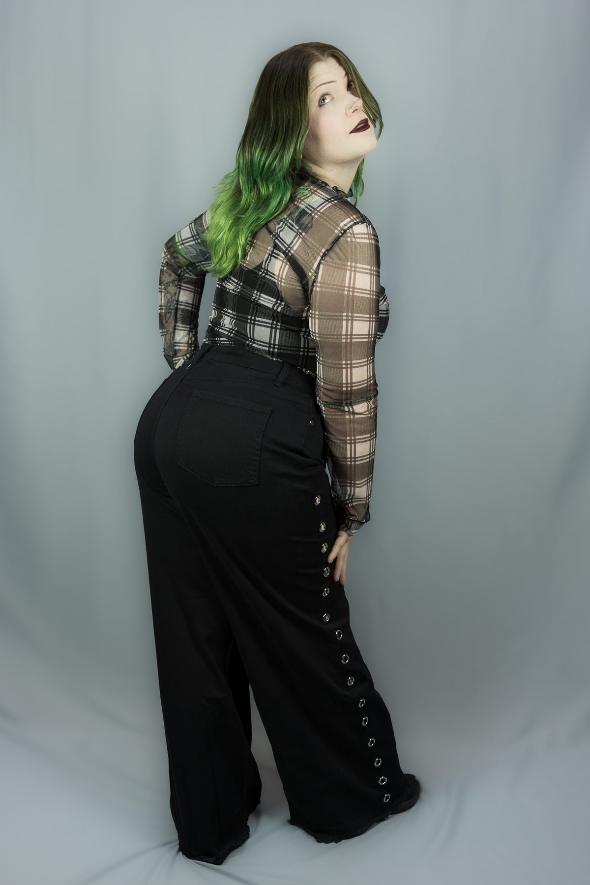 A model with green-tipped hair is shown from a rear three-quarter view, wearing a white and black plaid sheer long-sleeve shirt over a black top, paired with black wide-leg pants featuring side button details, against a light gray studio backdrop.