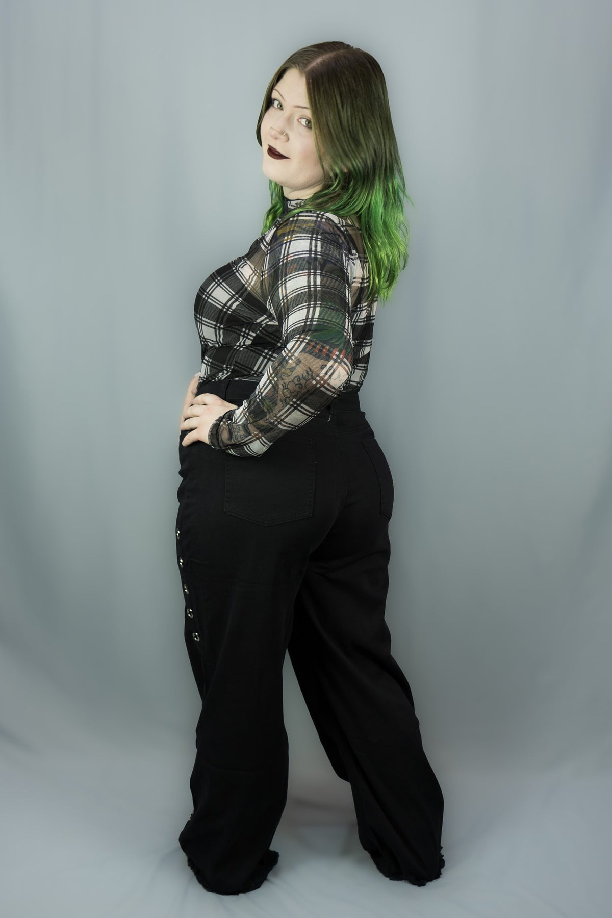 A model with green-tipped hair poses in a black and white plaid long-sleeve top and black wide-leg pants against a gray studio backdrop.