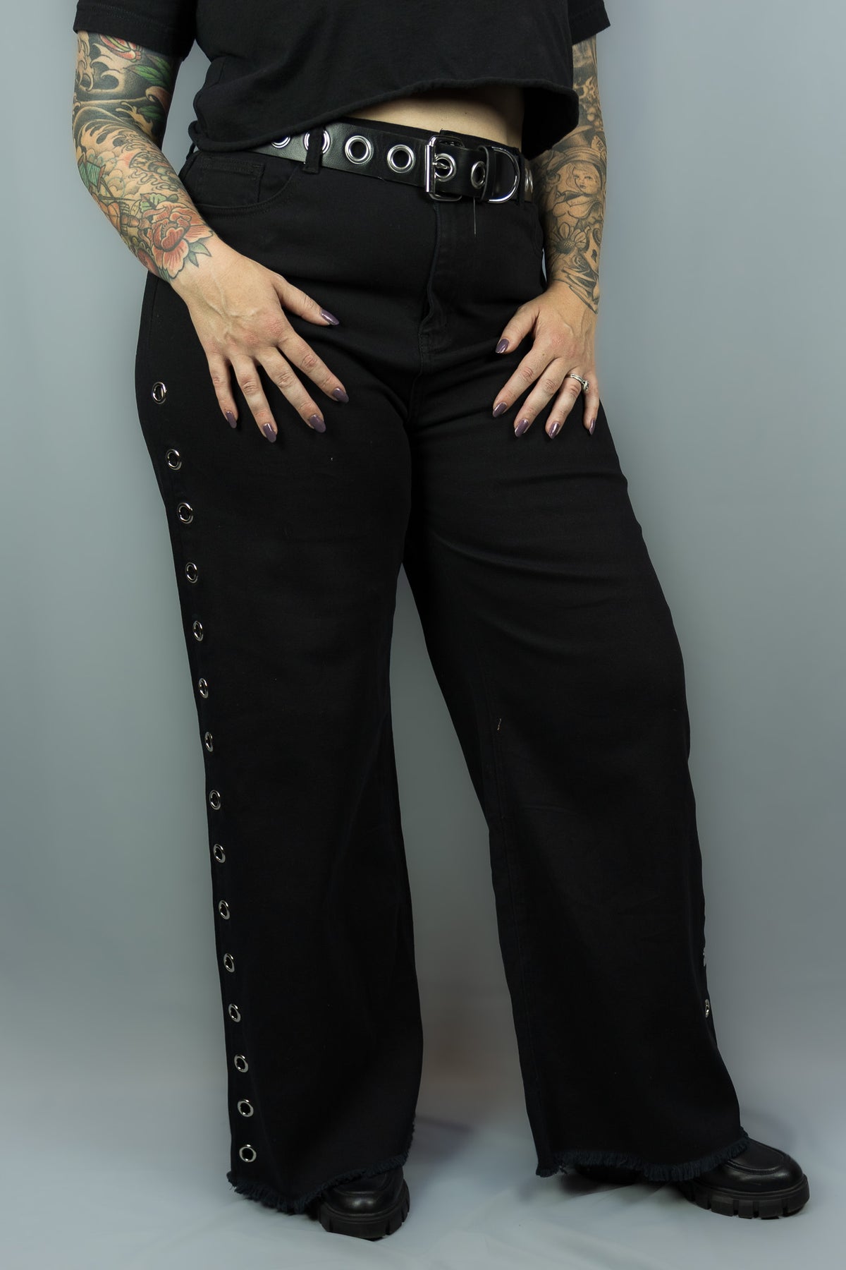 Black wide-leg pants with silver grommet details down the outer side of each leg, worn by a model with a black belt and black top against a gray studio background.