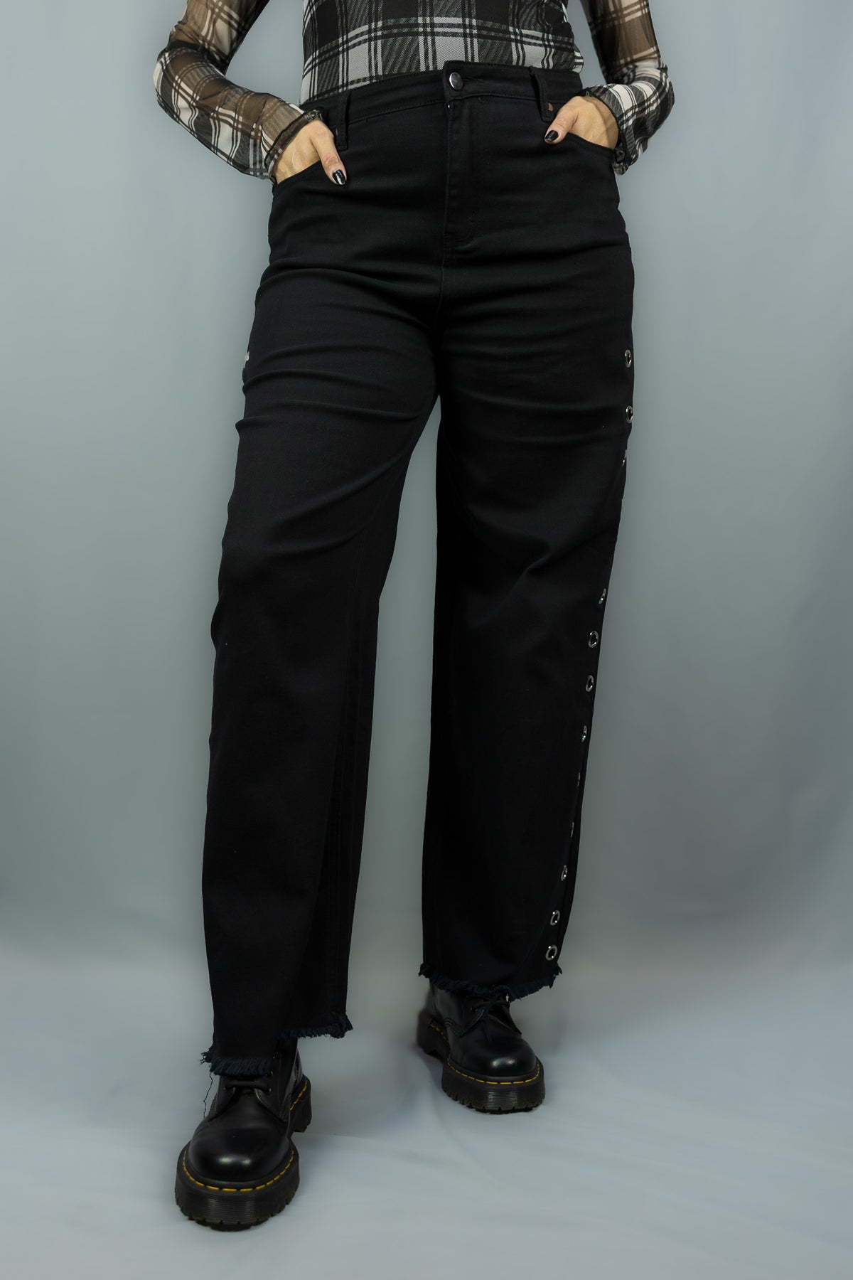 A person wearing black wide-leg jeans with side grommet details, paired with a plaid long-sleeve shirt and black platform boots, standing against a light gray studio backdrop.