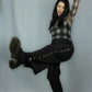A woman posing dynamically in a studio setting, wearing a black and white plaid long-sleeve top, black wide-leg pants with silver-tone studs, and black boots against a light gray backdrop.