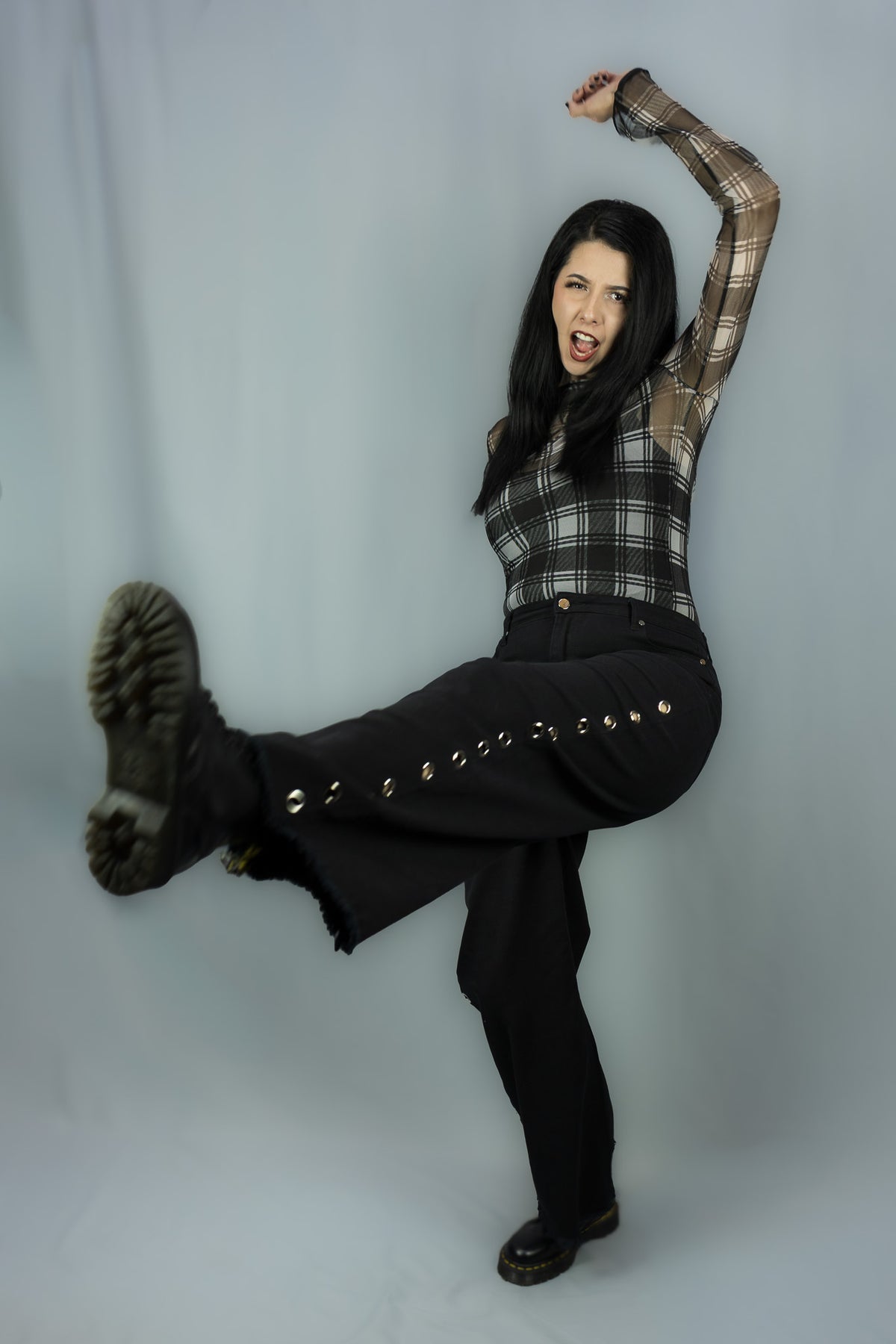 A woman posing dynamically in a studio setting, wearing a black and white plaid long-sleeve top, black wide-leg pants with silver-tone studs, and black boots against a light gray backdrop.