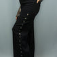 A person wearing black wide-leg jeans with a row of decorative metal eyelets down the side, paired with a white and black plaid long-sleeve top and black boots, against a light gray studio background.