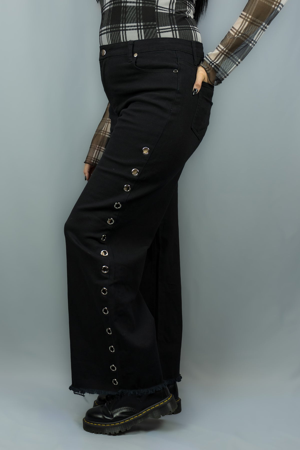 A person wearing black wide-leg jeans with a row of decorative metal eyelets down the side, paired with a white and black plaid long-sleeve top and black boots, against a light gray studio background.