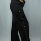 A person wearing black wide-leg pants with a vertical row of metal grommets down the outer side, paired with a plaid long-sleeve top and black boots, photographed against a light gray studio backdrop.