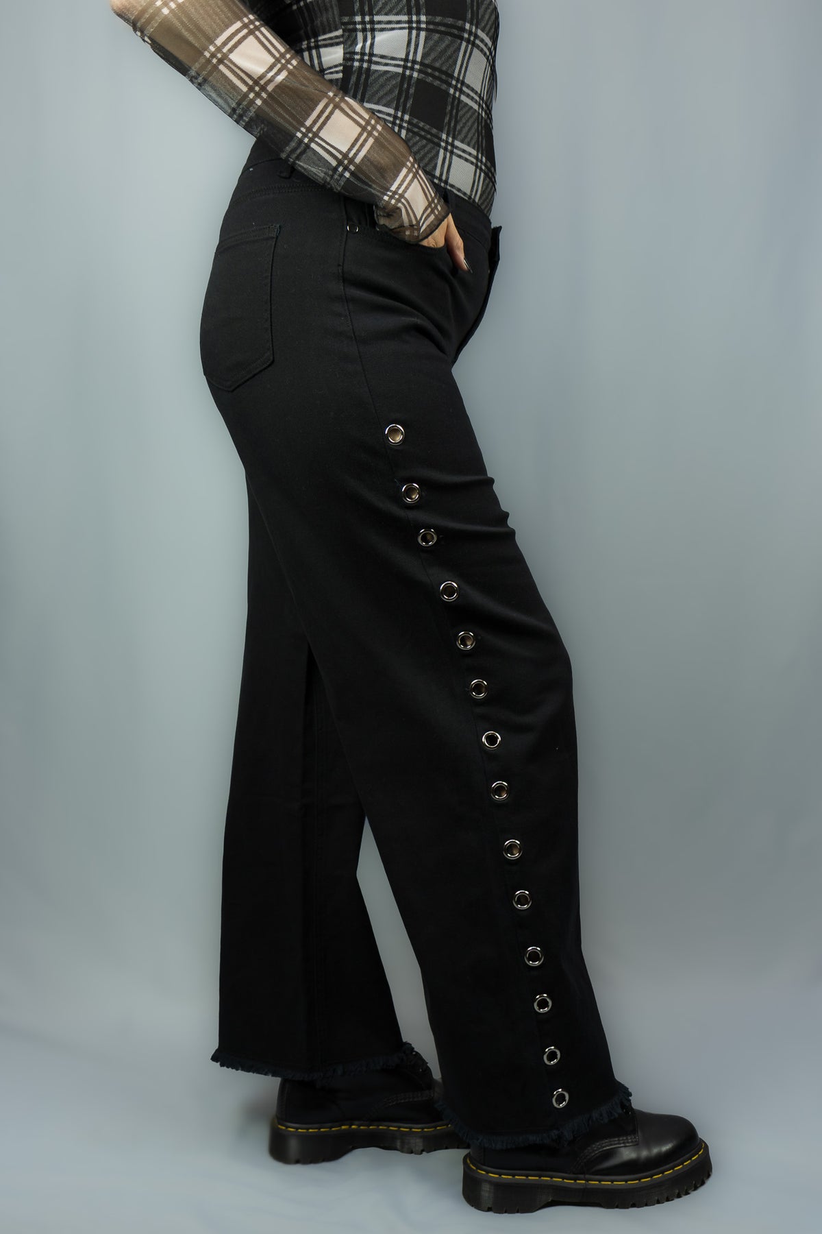A person wearing black wide-leg pants with a vertical row of metal grommets down the outer side, paired with a plaid long-sleeve top and black boots, photographed against a light gray studio backdrop.