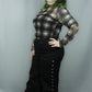 A woman modeling a black and white plaid long-sleeve top with sheer mesh sleeves and black wide-leg pants with decorative eyelets down the side, posed against a solid gray studio backdrop.
