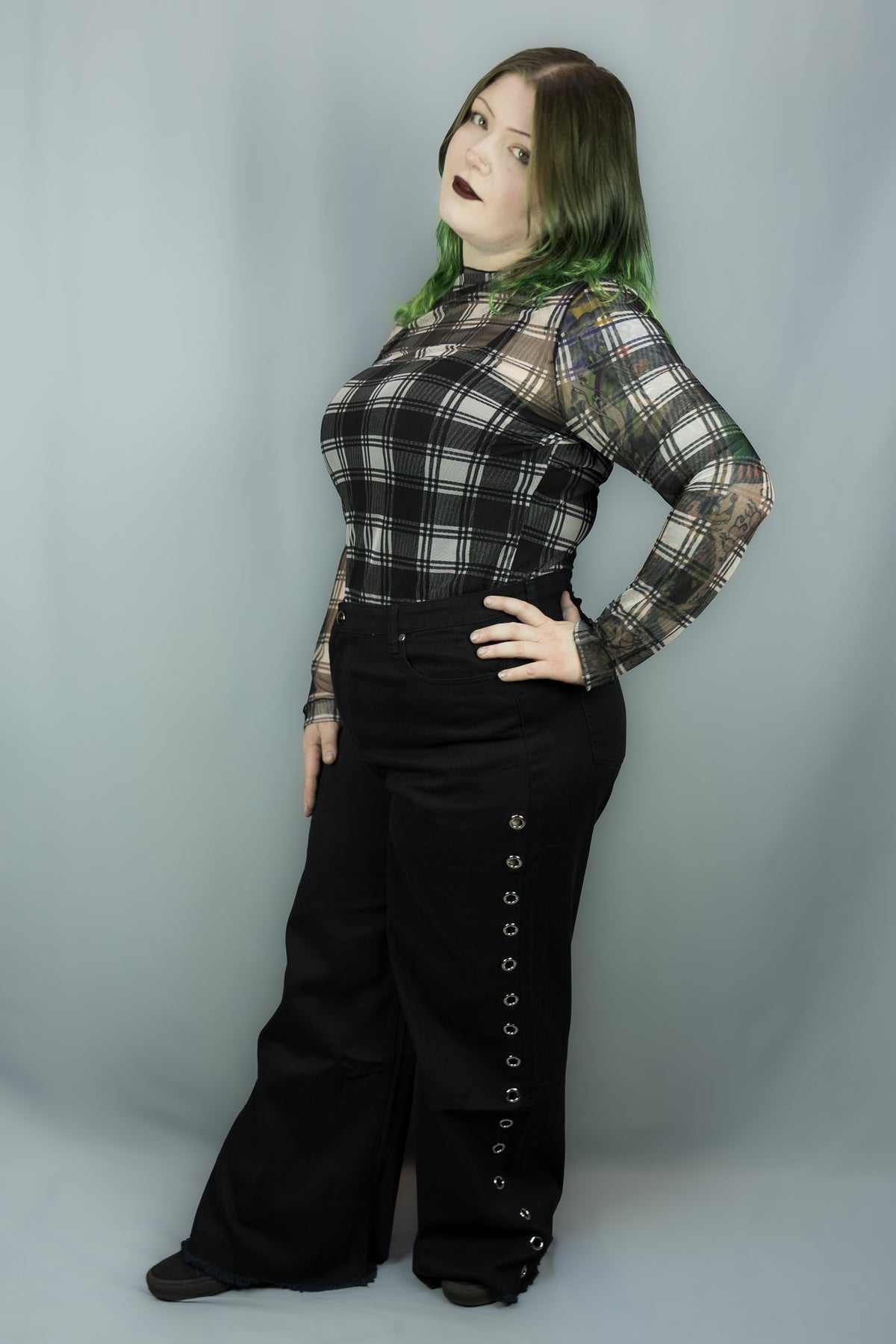 A woman modeling a black and white plaid long-sleeve top with sheer mesh sleeves and black wide-leg pants with decorative eyelets down the side, posed against a solid gray studio backdrop.