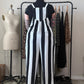 A person stands with their back to the camera wearing a black and white vertically striped jumpsuit over a black t-shirt, standing in front of a clothing rack in a room with a dress form and shelves.