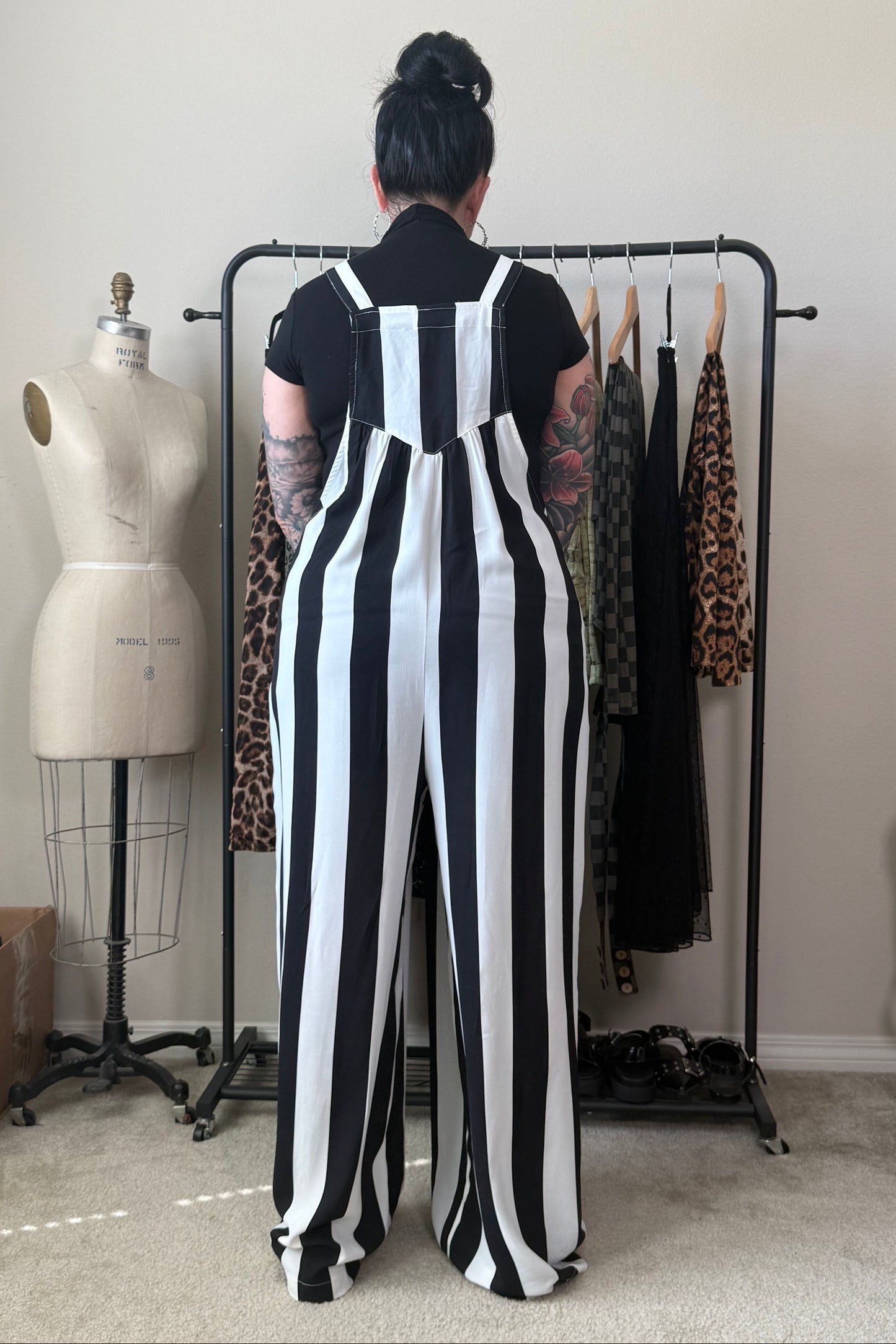 A person stands with their back to the camera wearing a black and white vertically striped jumpsuit over a black t-shirt, standing in front of a clothing rack in a room with a dress form and shelves.