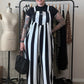 A person with short dark hair and visible arm tattoos stands indoors wearing a black-and-white vertically striped sleeveless jumpsuit over a black top. They are holding a black woven handbag, with a clothing rack, dress form, and room furniture visible in the background.