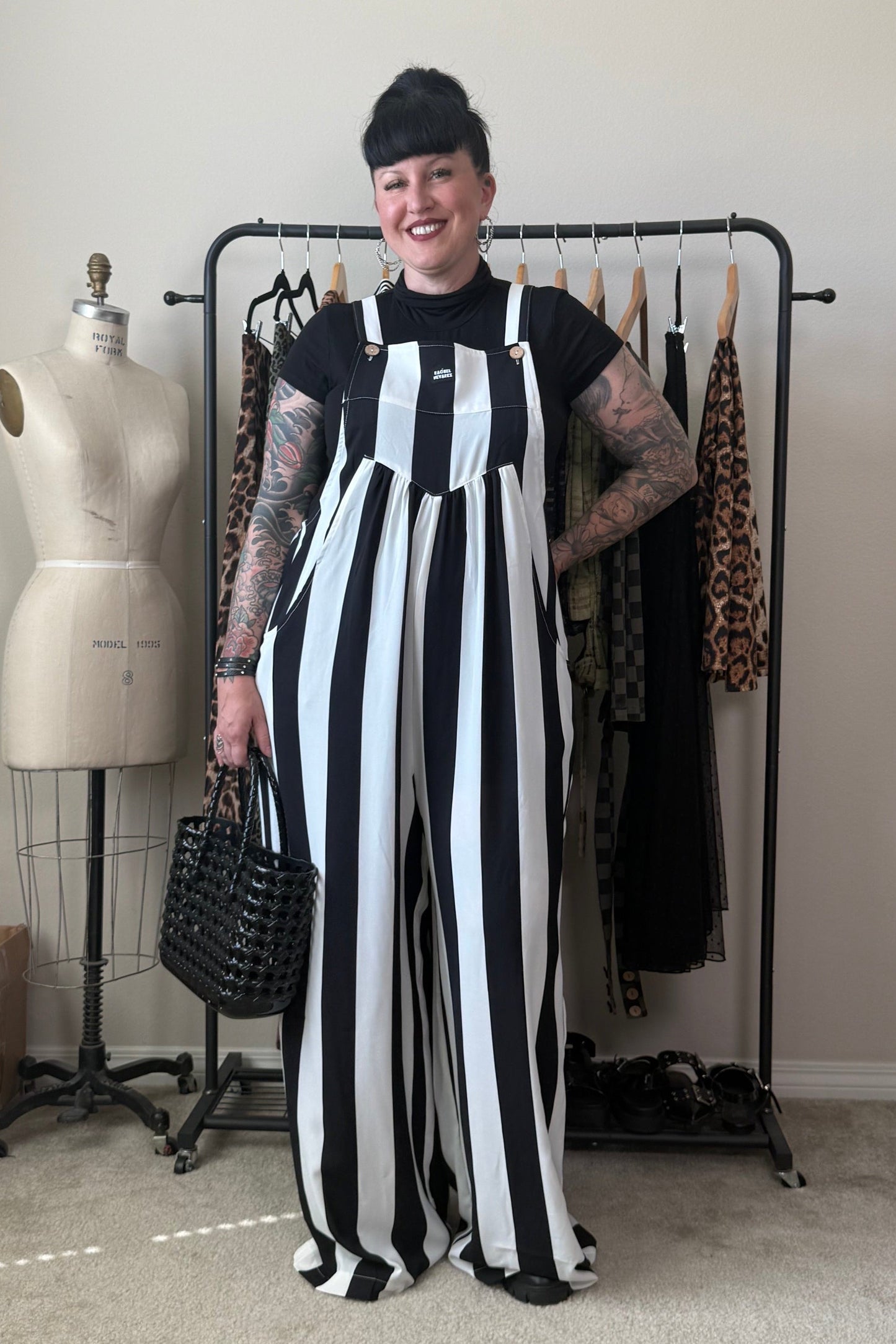 A person with short dark hair and visible arm tattoos stands indoors wearing a black-and-white vertically striped sleeveless jumpsuit over a black top. They are holding a black woven handbag, with a clothing rack, dress form, and room furniture visible in the background.