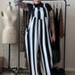 A person with short black hair and arm tattoos stands in a room wearing a black-and-white vertically striped wide-leg jumpsuit over a black top. A clothing rack with patterned garments, a dress form, and room furnishings are visible in the background.