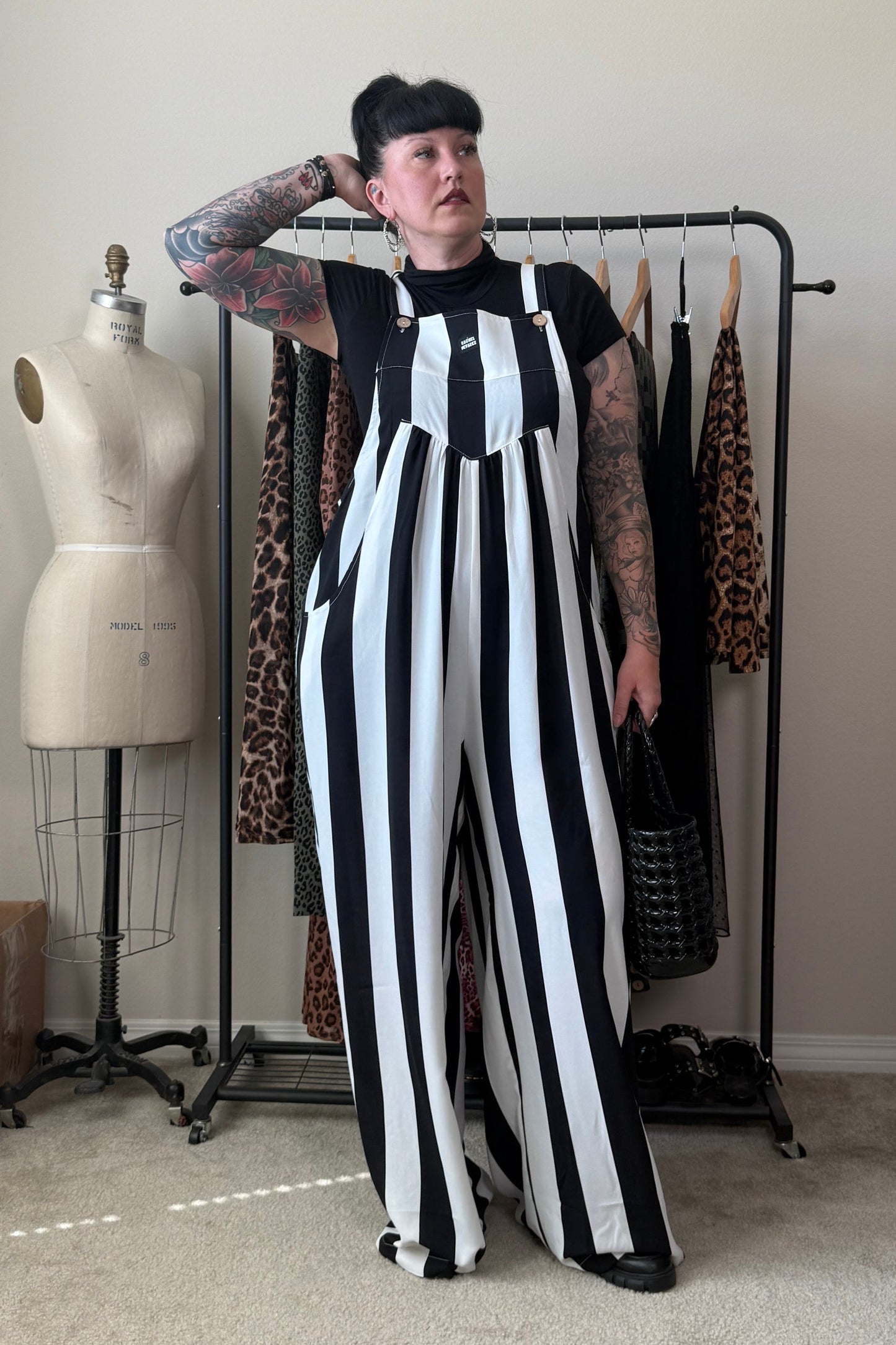 A person with short black hair and arm tattoos stands in a room wearing a black-and-white vertically striped wide-leg jumpsuit over a black top. A clothing rack with patterned garments, a dress form, and room furnishings are visible in the background.