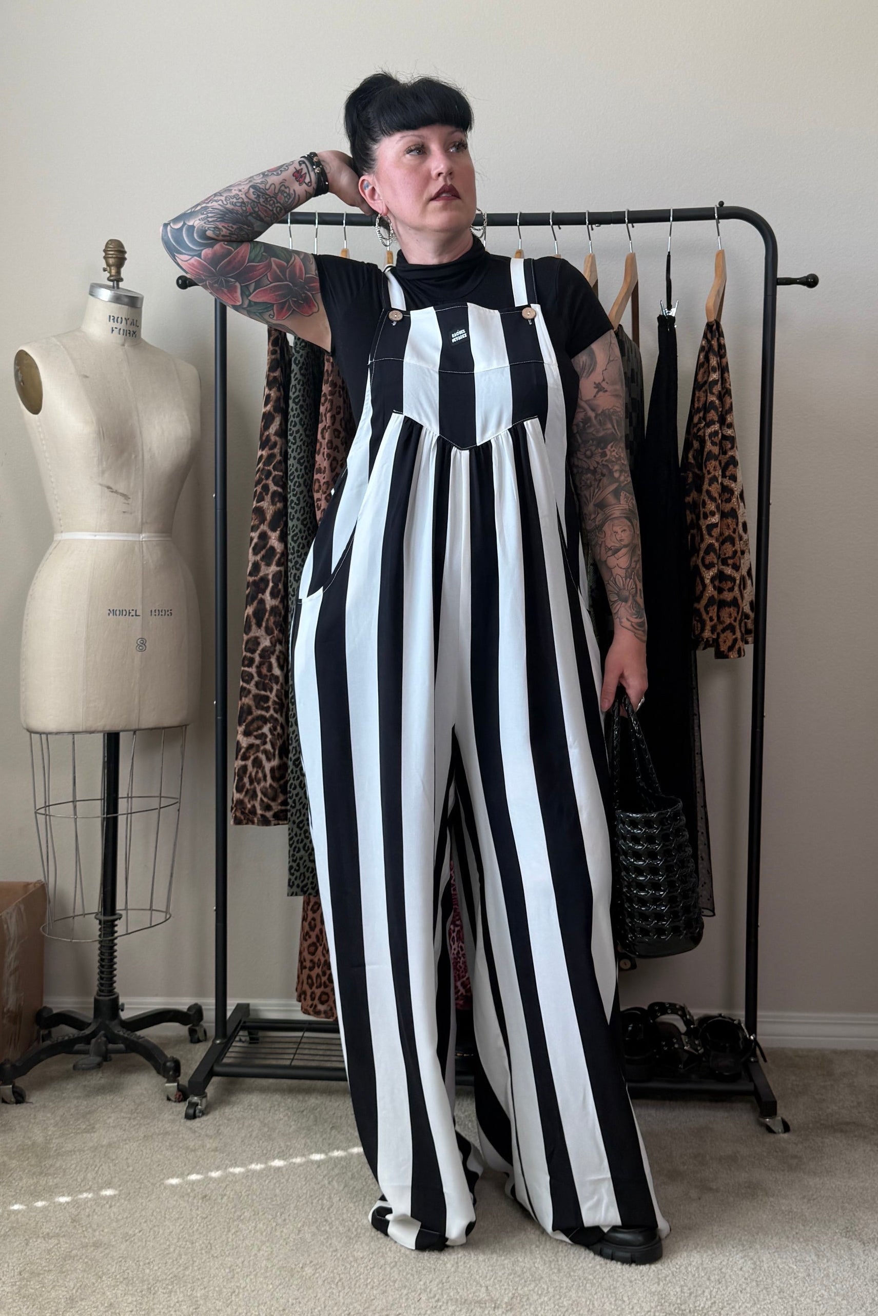 A person with short black hair and arm tattoos stands in a room wearing a black-and-white vertically striped wide-leg jumpsuit over a black top. A clothing rack with patterned garments, a dress form, and room furnishings are visible in the background.