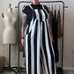 A person stands indoors wearing a black-and-white vertically striped wide-leg jumpsuit over a black short-sleeve top. A dress form, clothing rack, and room furniture are visible in the background.