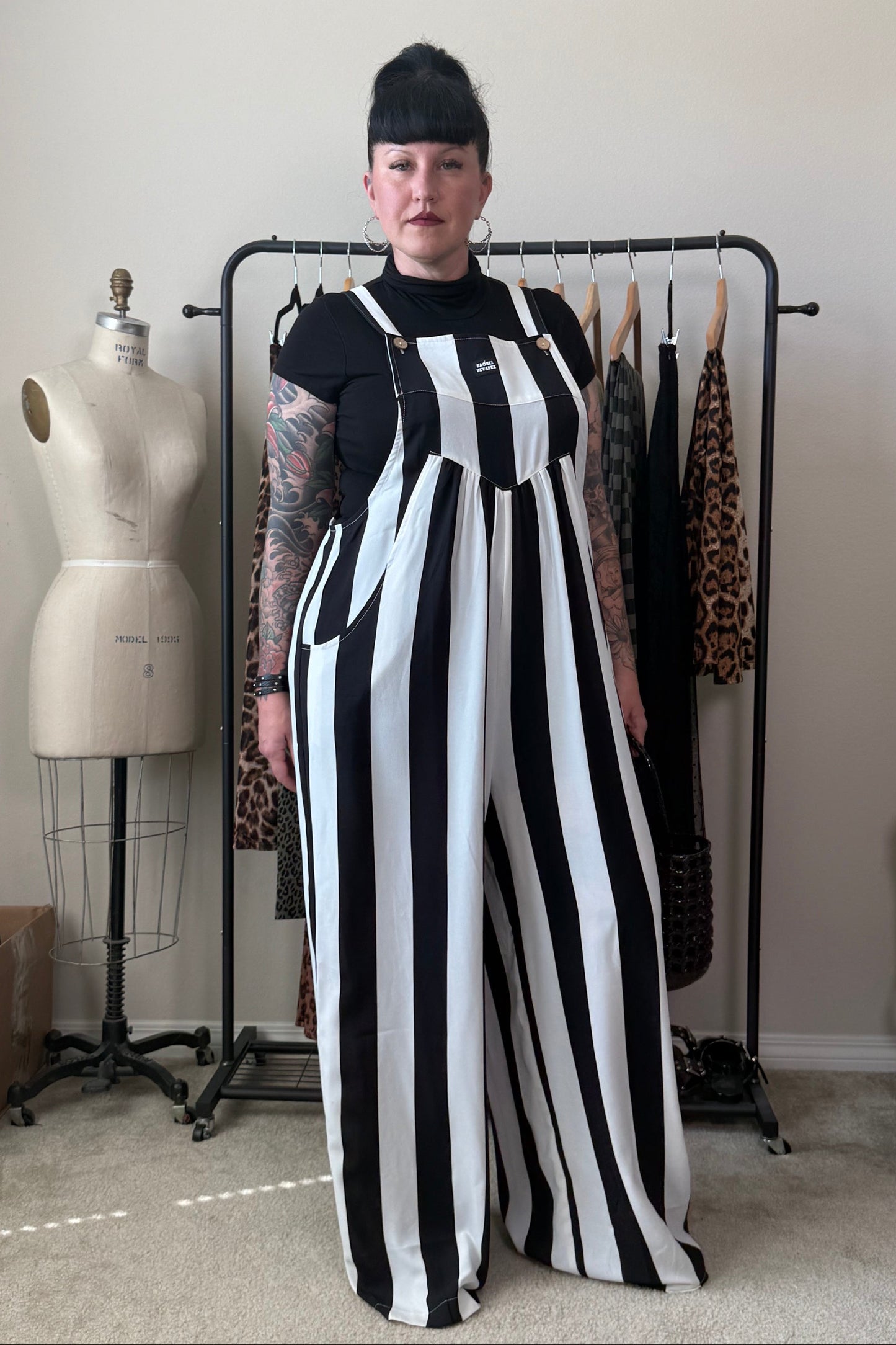 A person stands indoors wearing a black-and-white vertically striped wide-leg jumpsuit over a black short-sleeve top. A dress form, clothing rack, and room furniture are visible in the background.
