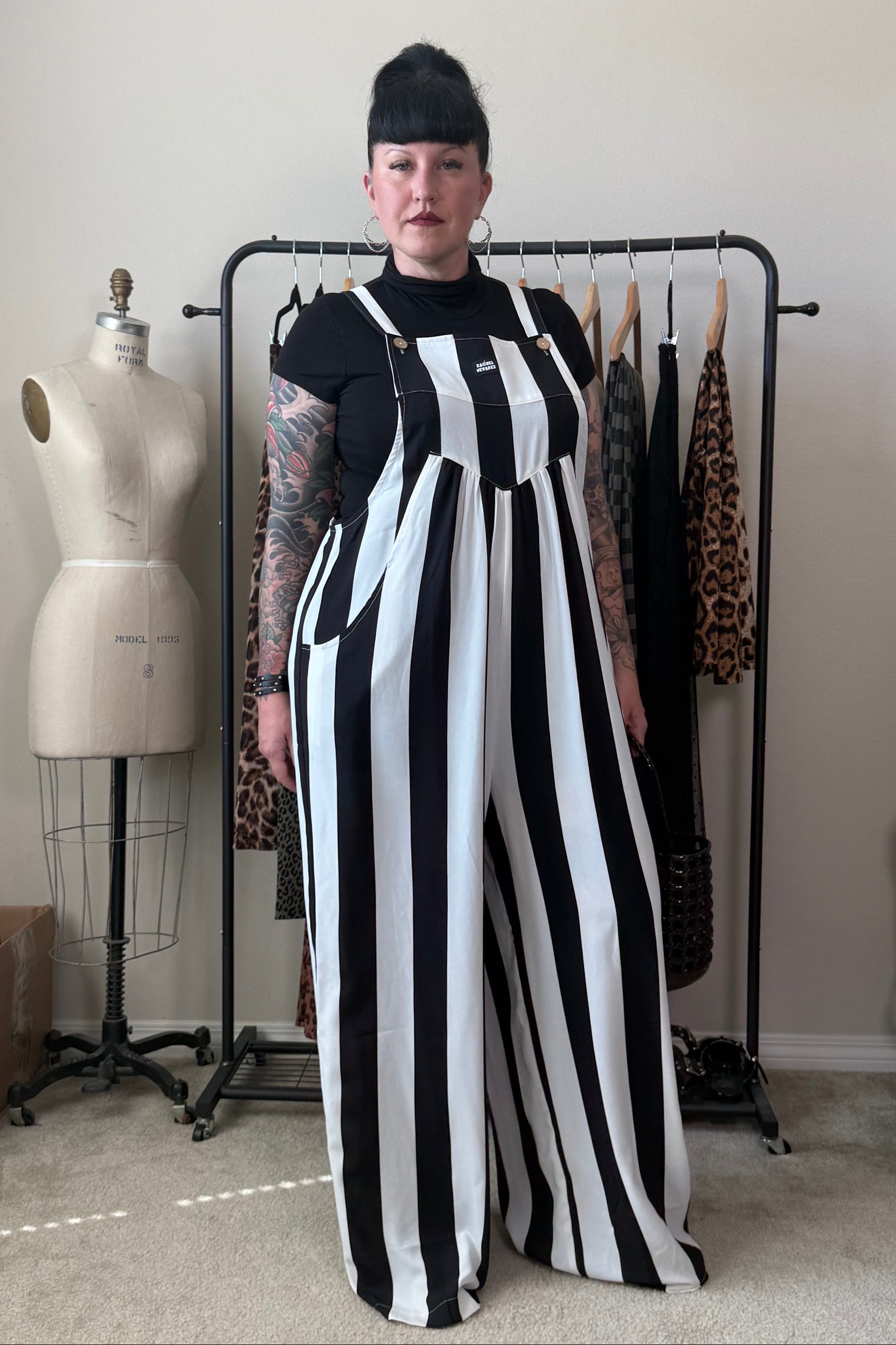 A person stands indoors wearing a black-and-white vertically striped wide-leg jumpsuit over a black short-sleeve top. A dress form, clothing rack, and room furniture are visible in the background.