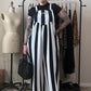 A person stands in a room wearing a black and white vertically striped jumpsuit over a black t-shirt, holding a black woven handbag. A dress form, clothing rack, and room elements are visible in the background.